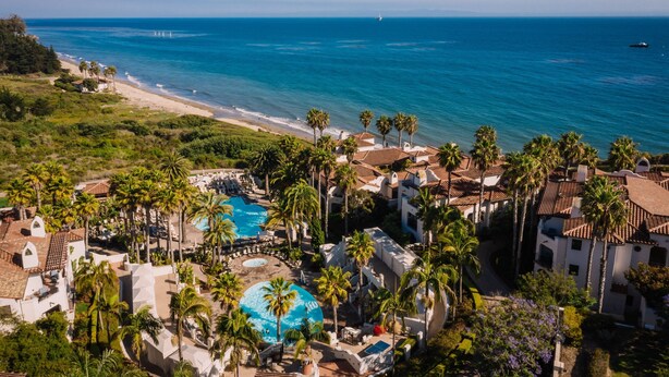 An aerial view of a resort with a large swimming pool, palm trees, and a beach.