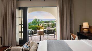 Une chambre d’hôtel avec un lit, un canapé, une lampe et une vue sur une terrasse extérieure aménagée avec des chaises et un foyer.