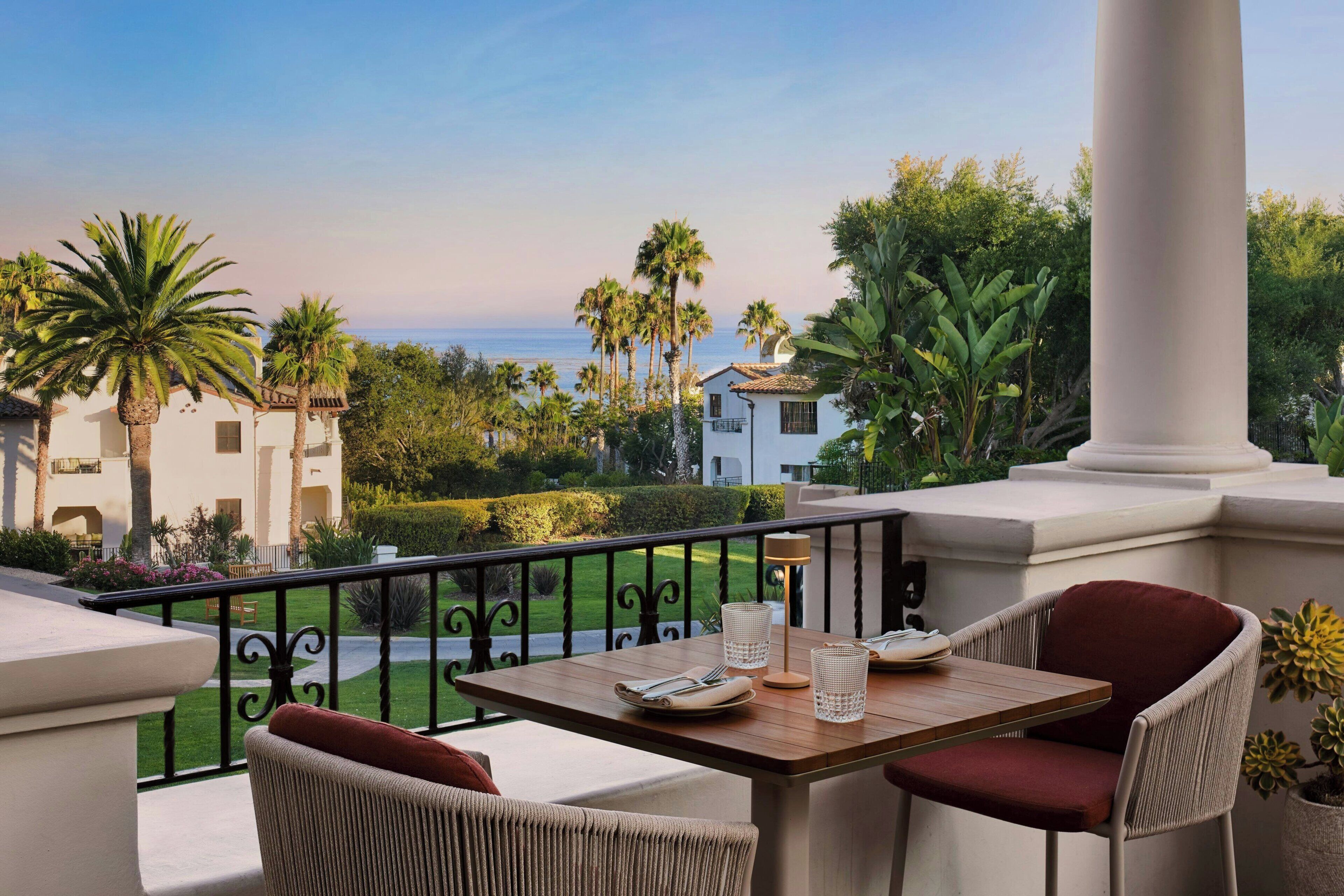 Un balcon avec une table et des chaises, donnant sur un jardin aménagé et offrant une vue sur la mer.