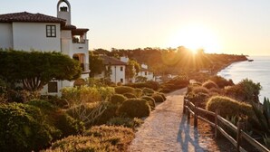 A coastal property with a pathway leading to a white building with a bell tower, surrounded by lush greenery and overlooking the sea.