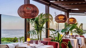 A restaurant with tables set for dining, large windows offering a view of the sea, and wicker hanging lights.