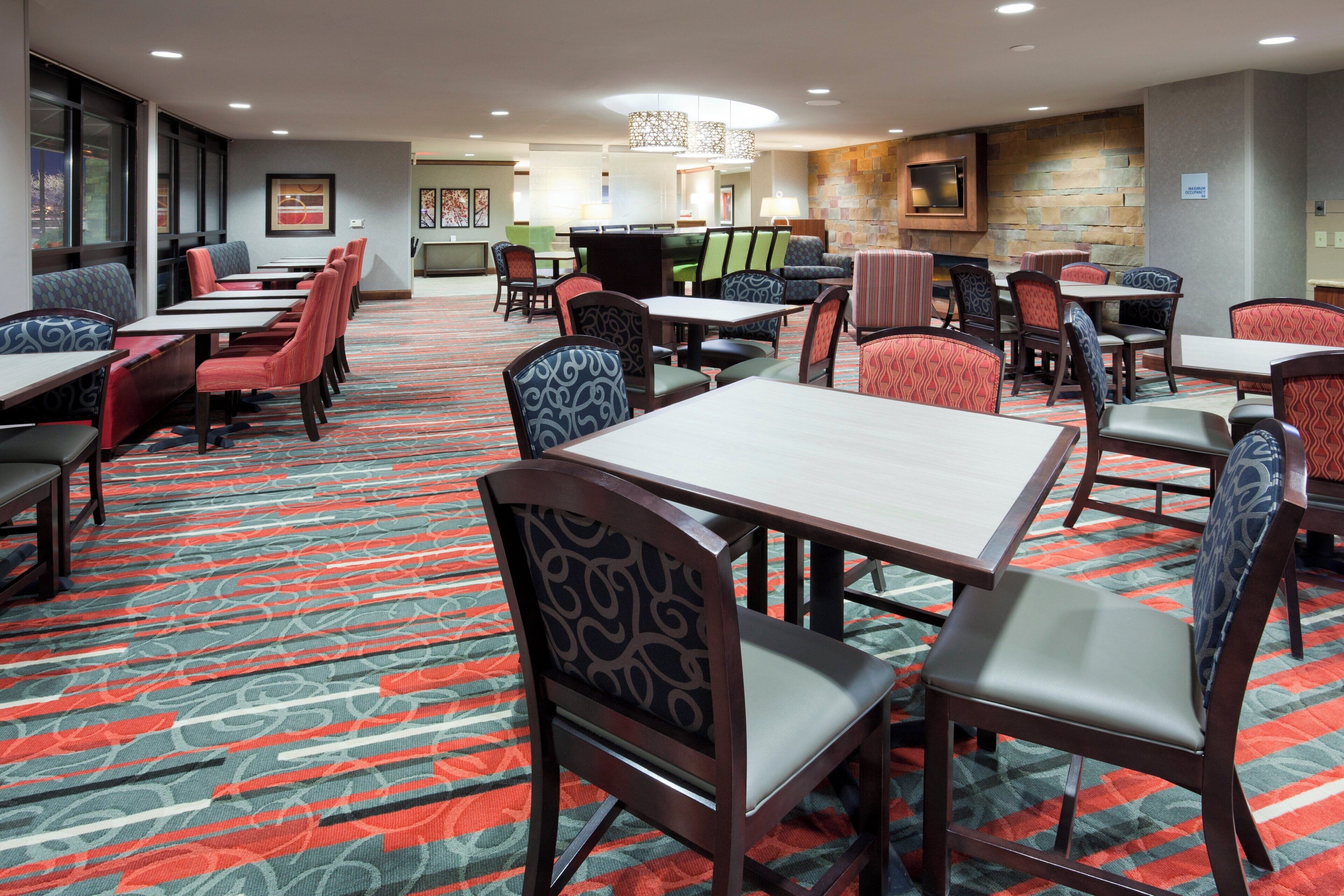 A hotel dining area with multiple tables and chairs, a patterned carpet, and a stone wall with a fireplace.