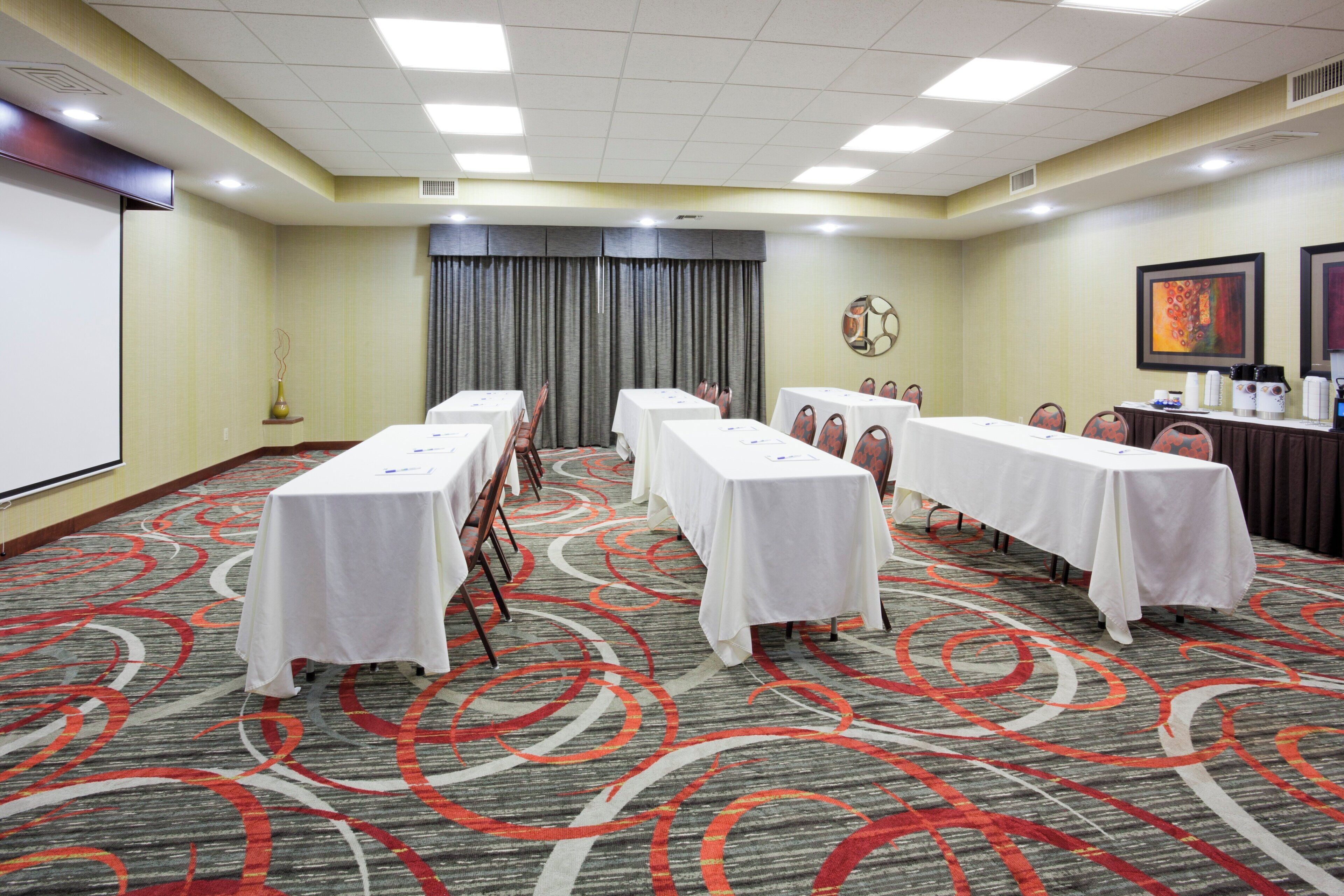 A conference room with multiple tables, chairs, a projector screen, and a carpeted floor.