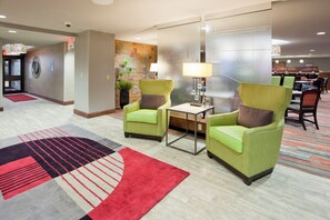 Un área de recepción con dos sillones verdes, una mesita con una lámpara, un tapete rojo y negro, y una pared con diseño de piedra.