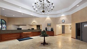 Un vestíbulo de hotel con una mesa de recepción, un reloj, una araña y una pequeña mesa con una planta.