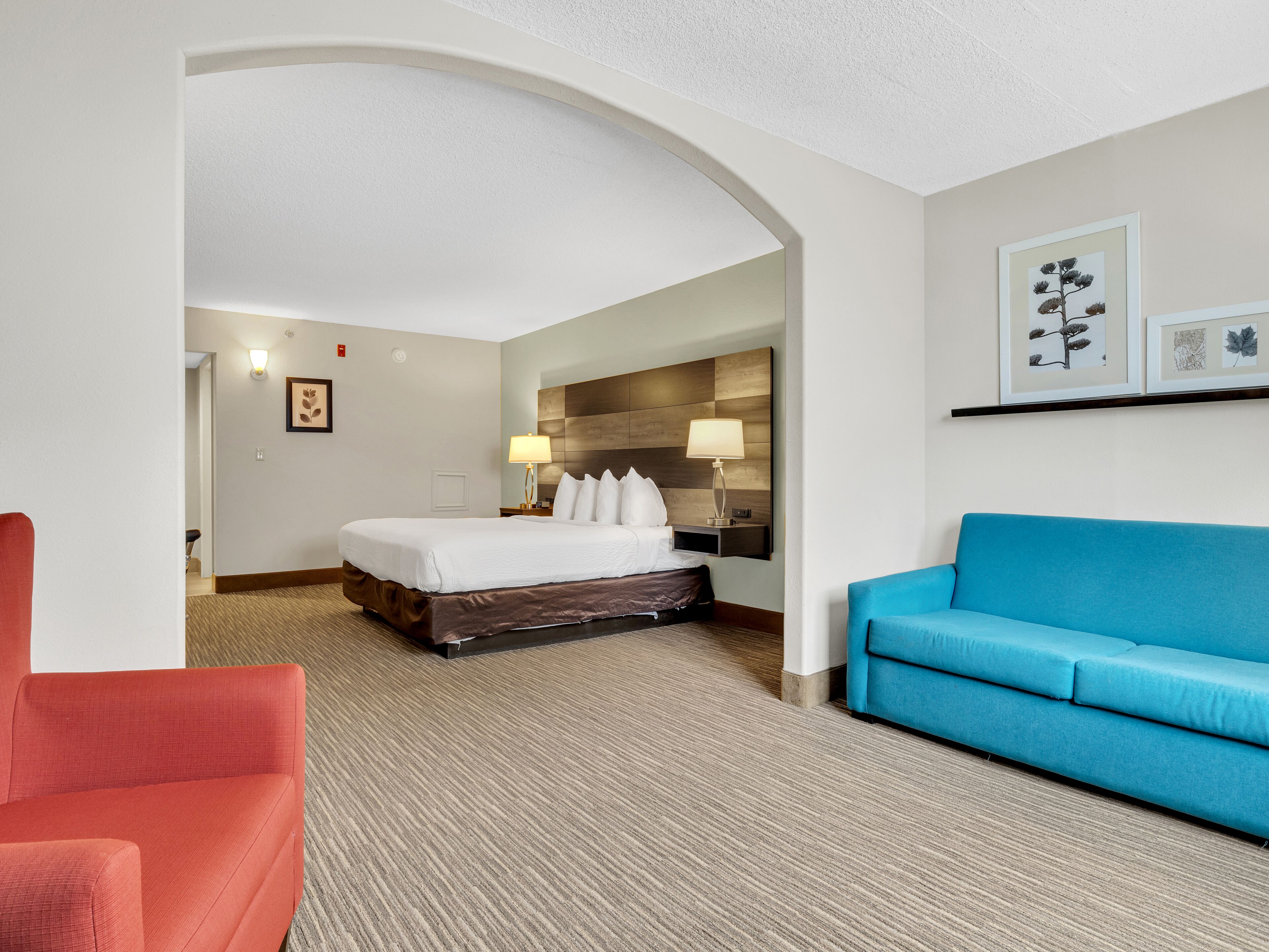 Habitación de hotel con cama, sofá azul, silla roja y un pasillo que conduce a otra habitación.