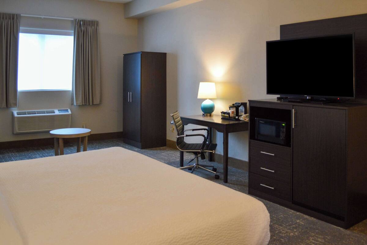 A hotel room with a large bed, a desk with a lamp, a television, a wardrobe, and a window with curtains.
