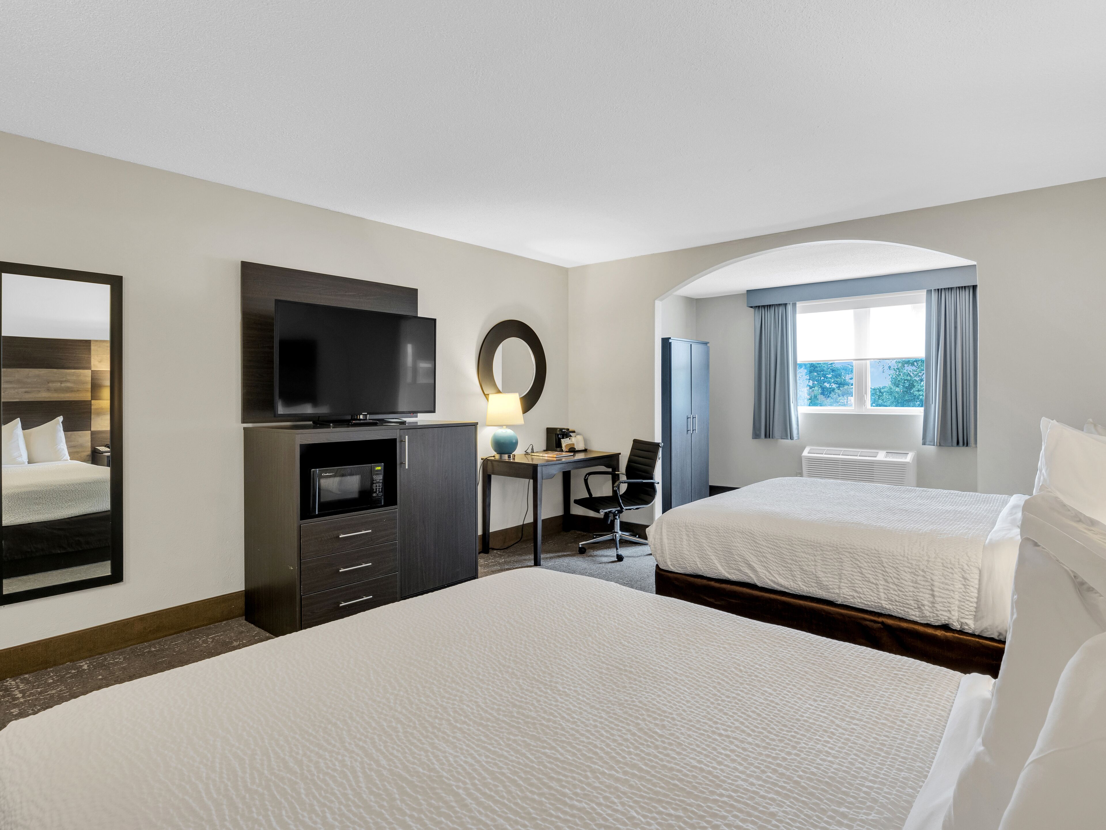 A hotel room with two beds, a desk with a chair, a television, a microwave, and a large window with curtains.