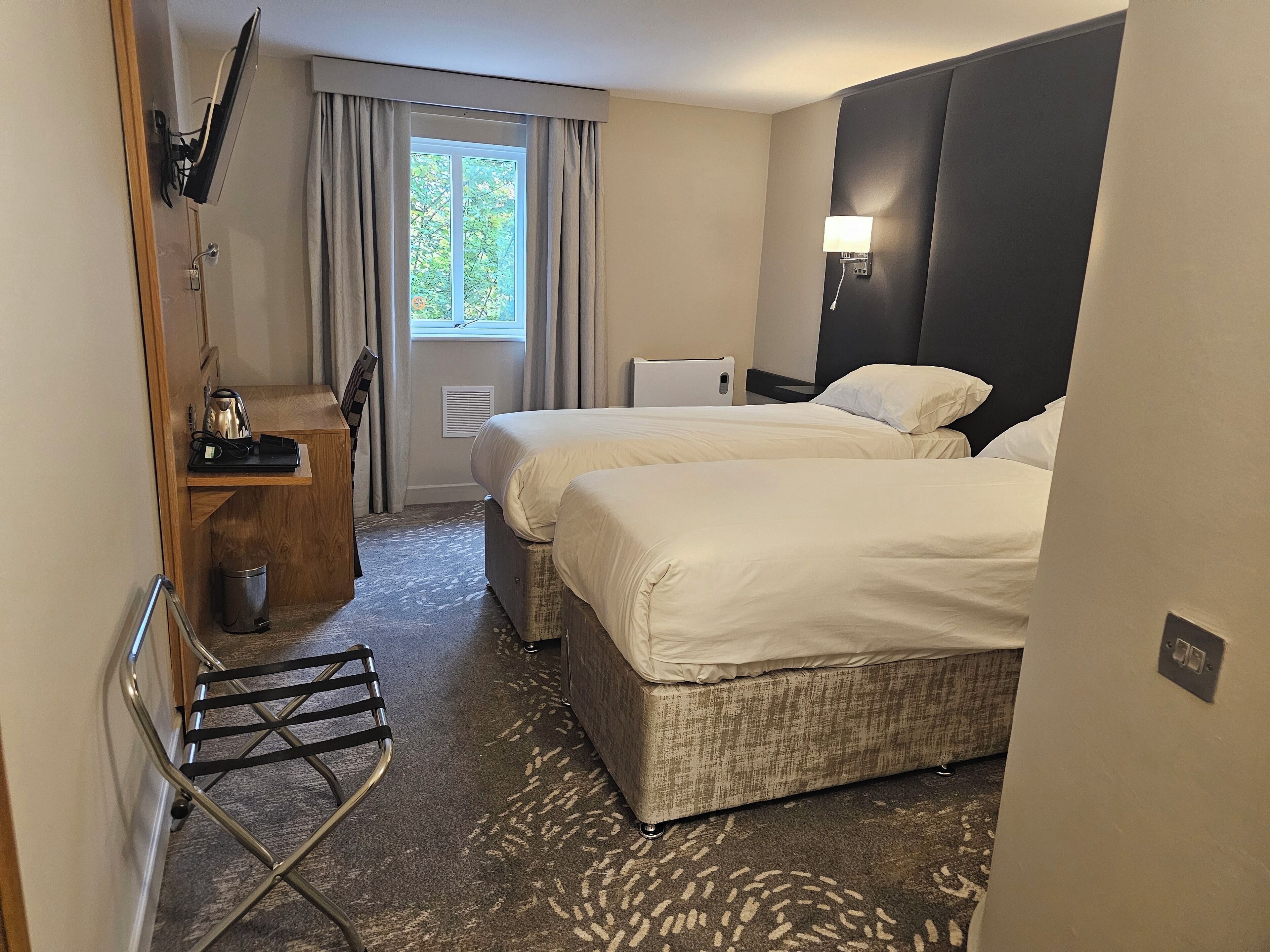 A hotel room with two beds, a desk with a kettle, a lamp, and a window with curtains.