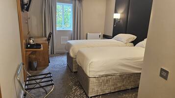 A hotel room with two beds, a desk with a kettle, a lamp, and a window with curtains.