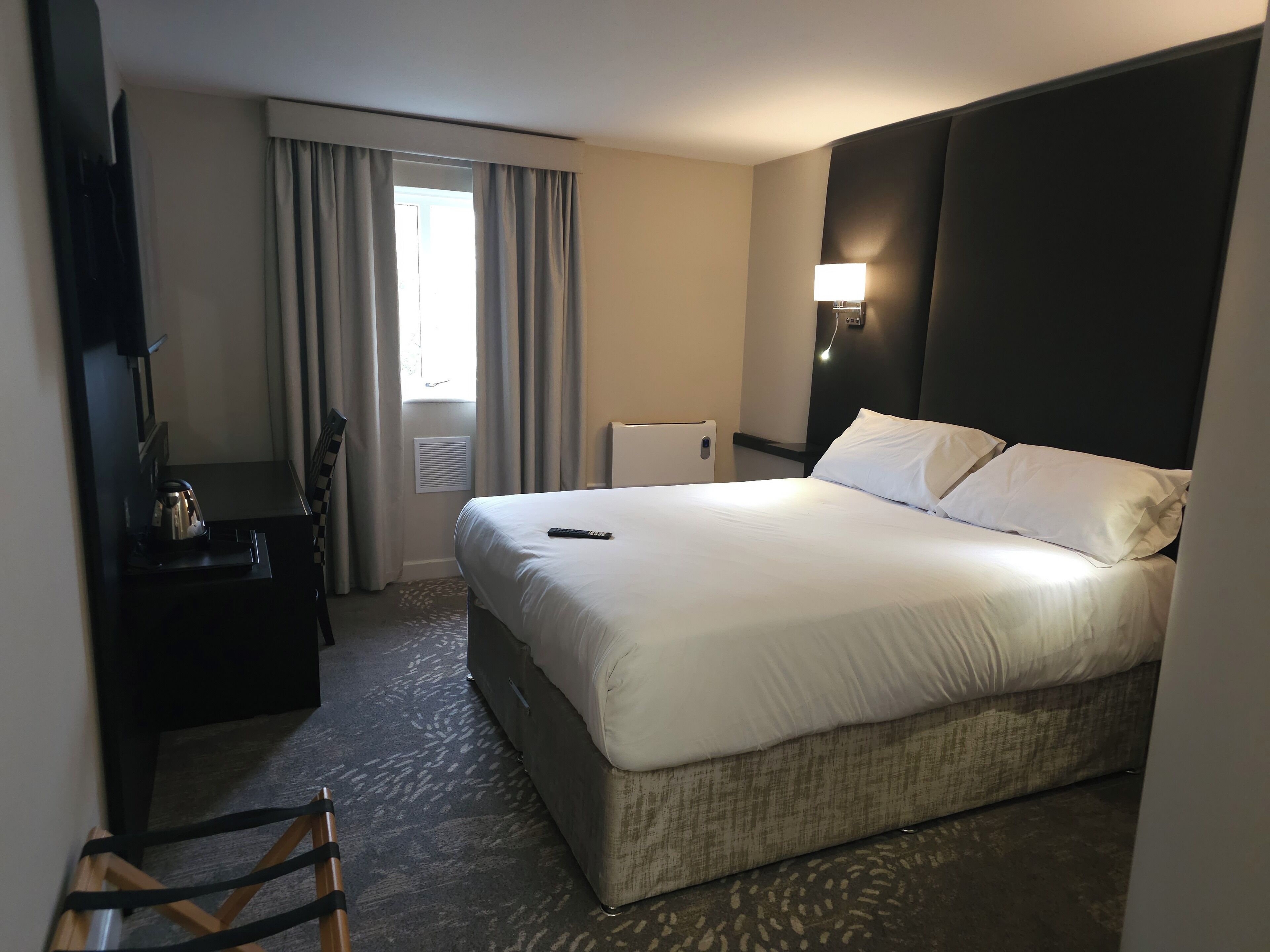 Ein Hotelzimmer mit einem großen Bett, einem Nachttisch mit Wasserkocher, einer Lampe und Blick ins Freie durch das Fenster.