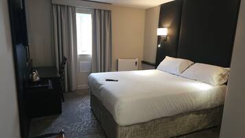 Ein Hotelzimmer mit einem großen Bett, einem Nachttisch mit Wasserkocher, einer Lampe und Blick ins Freie durch das Fenster.