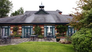 A building with a brick facade, green shutters, and a tiled roof. There are outdoor tables and chairs, and a well-maintained lawn.