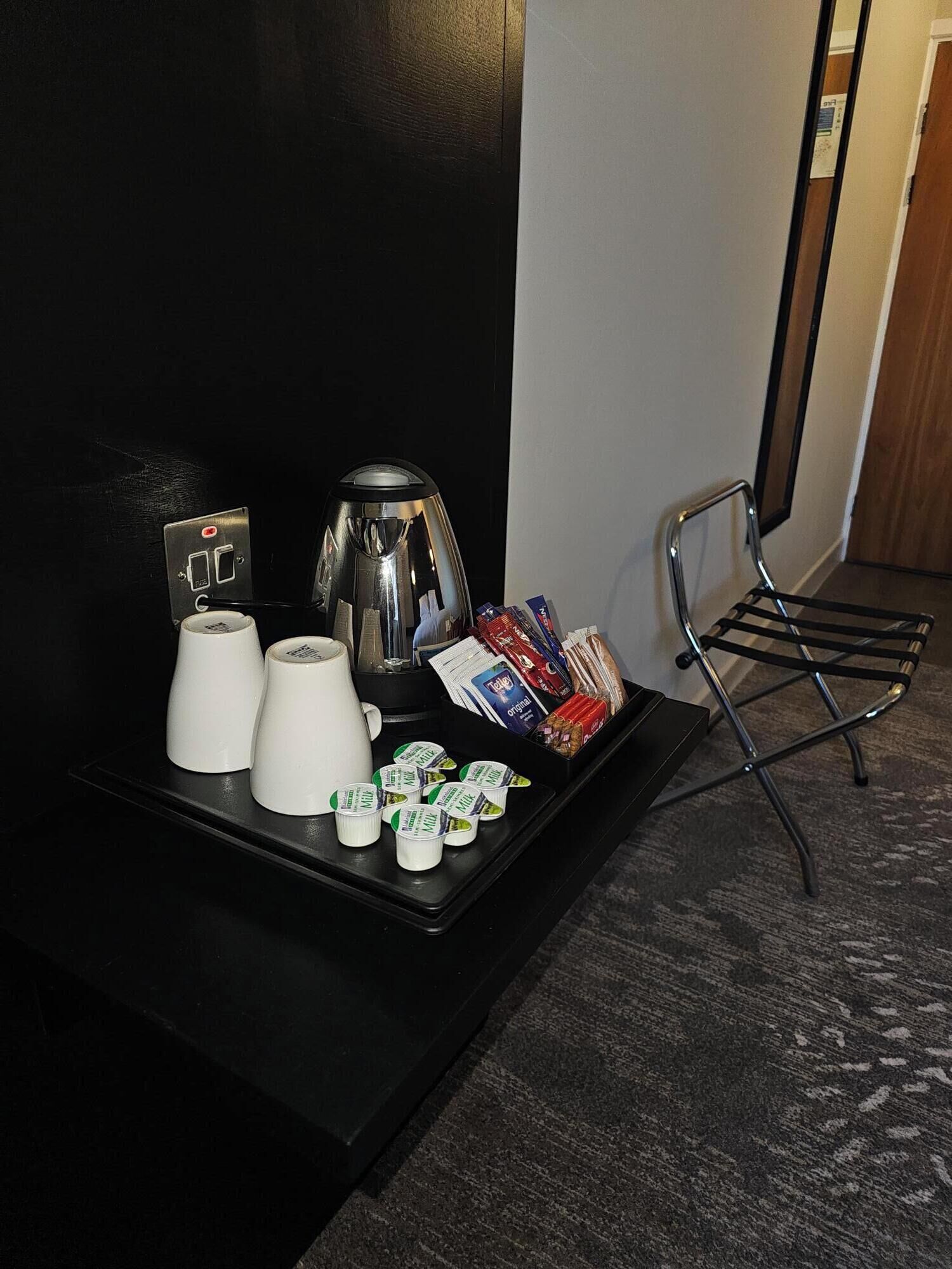 A hotel room with a black tray table holding a coffee maker, two white cups, a small refrigerator, and a tray of snacks.
