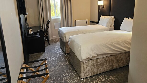 A hotel room with two beds, a desk, a chair, a lamp, and a window with curtains.