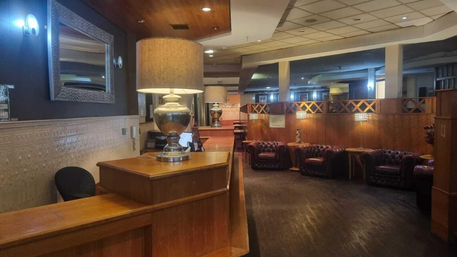 A reception area with a wooden counter, a large mirror, and a lamp. There are leather chairs and a wooden partition in the background.