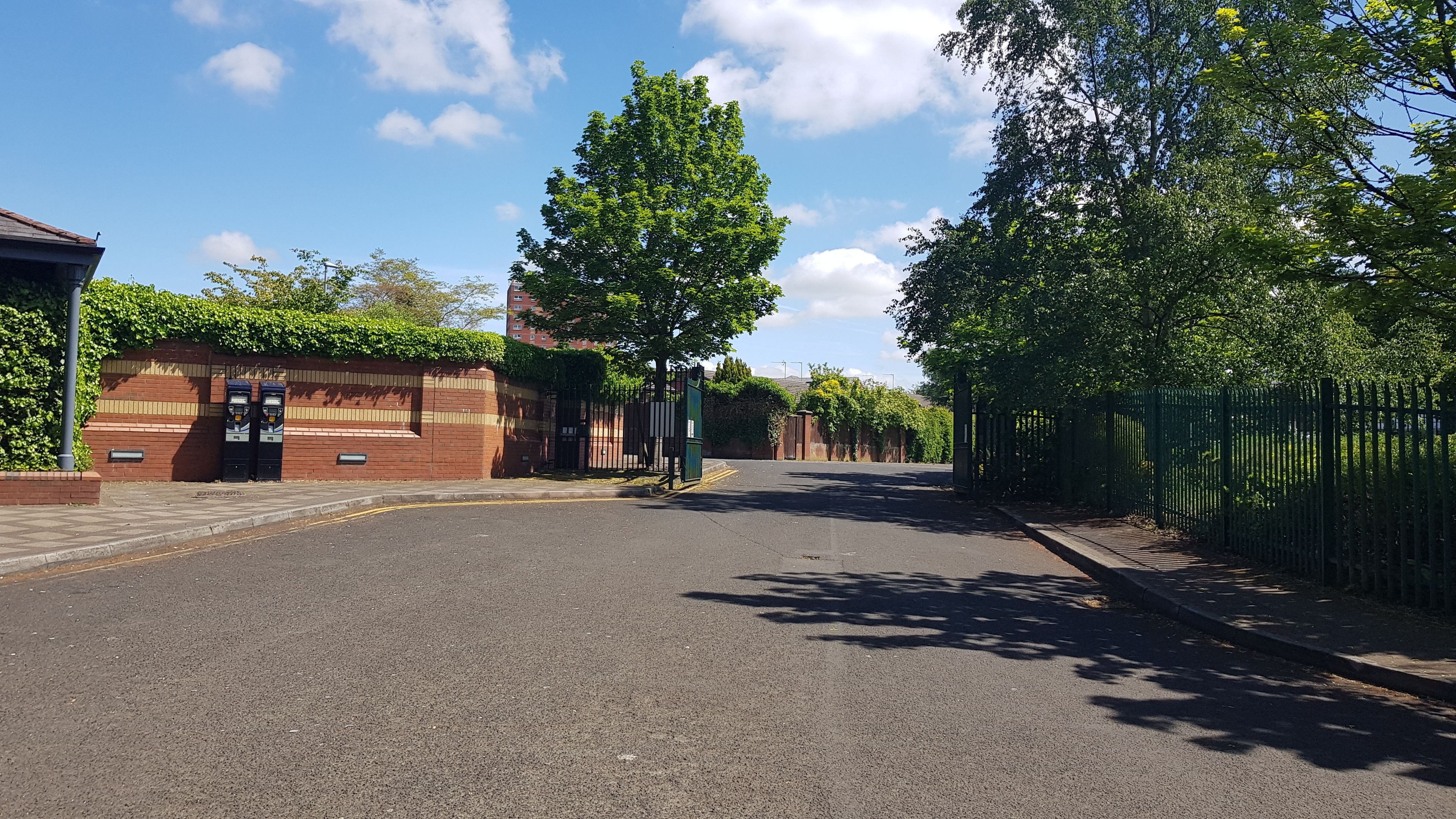 A quiet street with a brick building, a gated entrance, and a green fence.