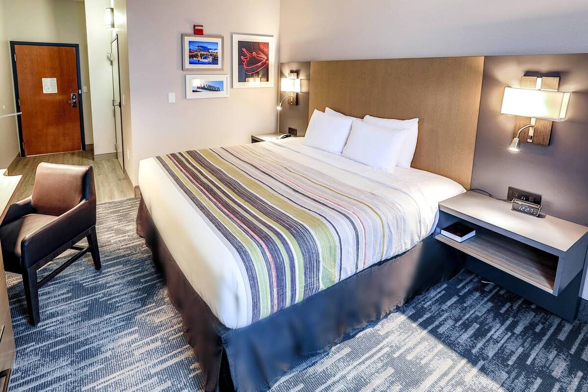 A hotel room with a large bed, a chair, a nightstand with a lamp, and a wall clock.