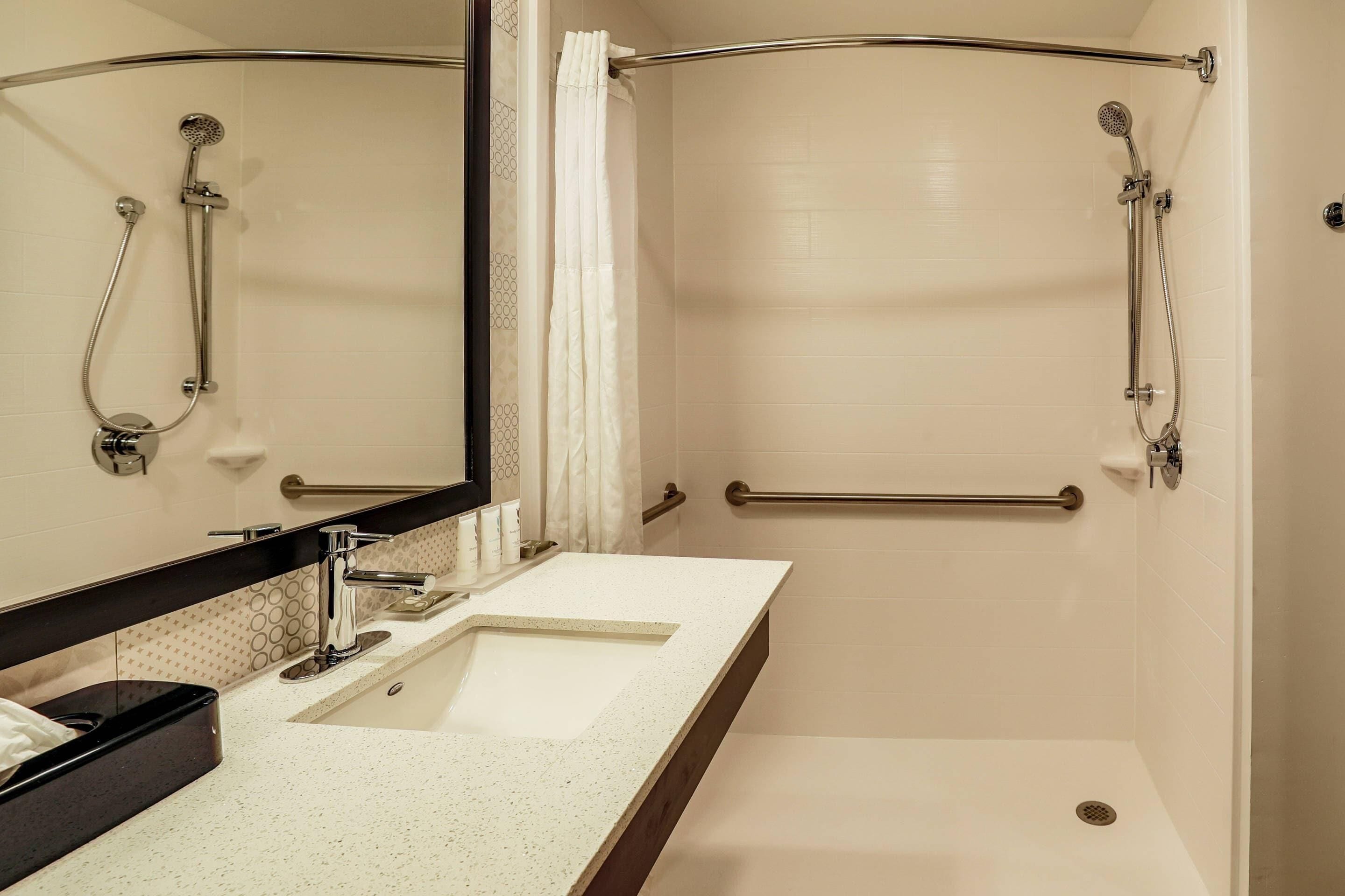 Une salle de bain moderne avec un lavabo blanc, un grand miroir, une douche équipée d’un pommeau de douche à main et d’une barre d’appui.