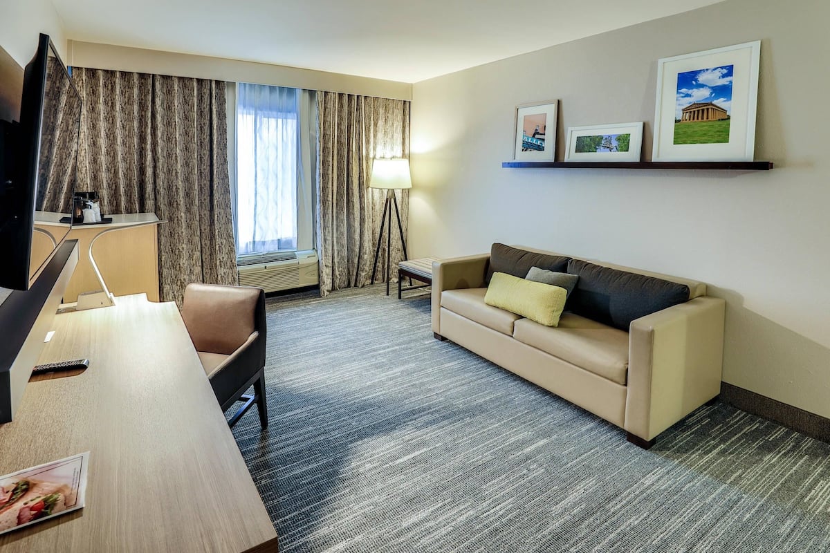 A hotel room with a desk, a sofa, a TV, a lamp, and framed pictures on the wall.