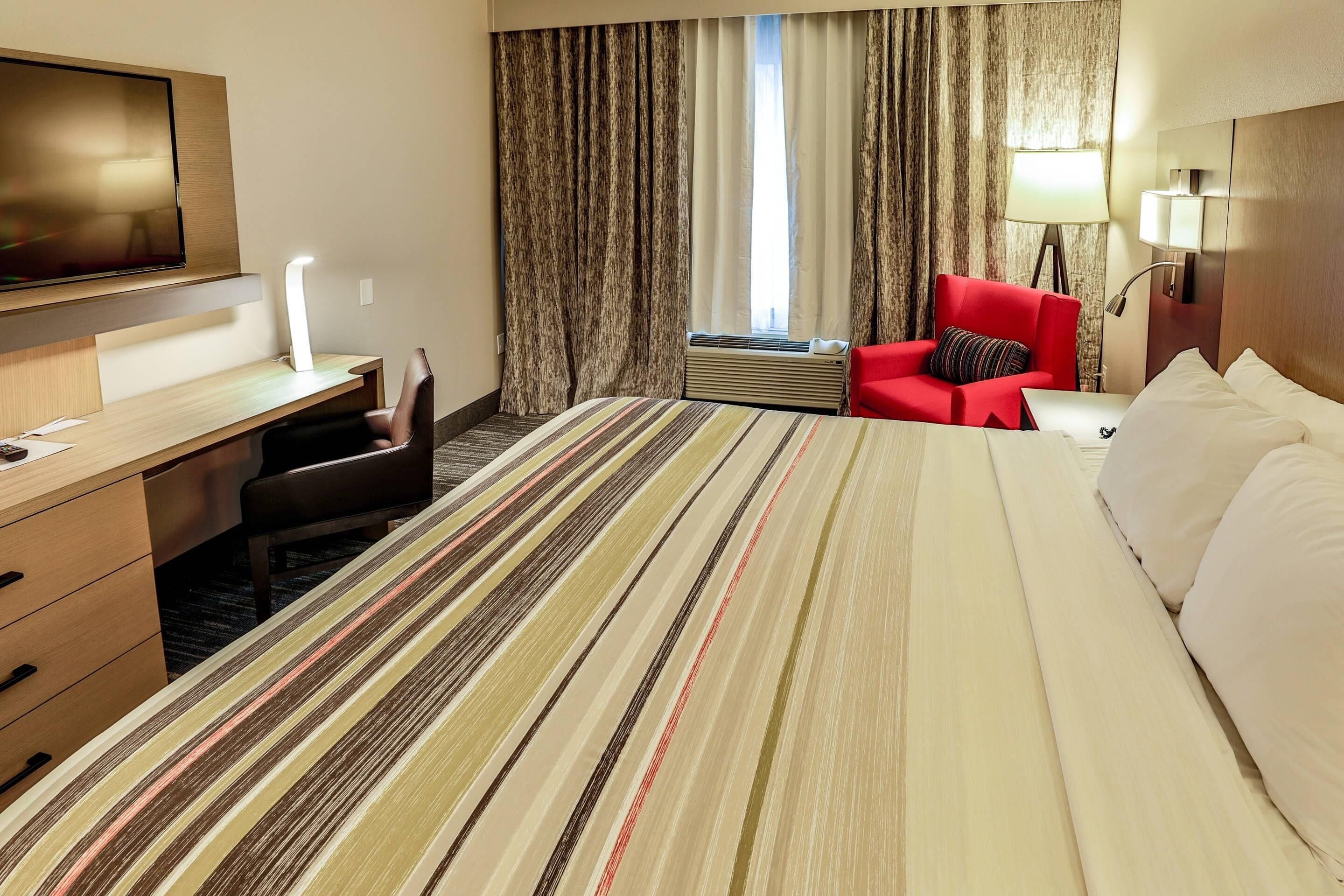 Habitación de hotel con cama, escritorio con silla, televisor, sillón rojo y lámpara.