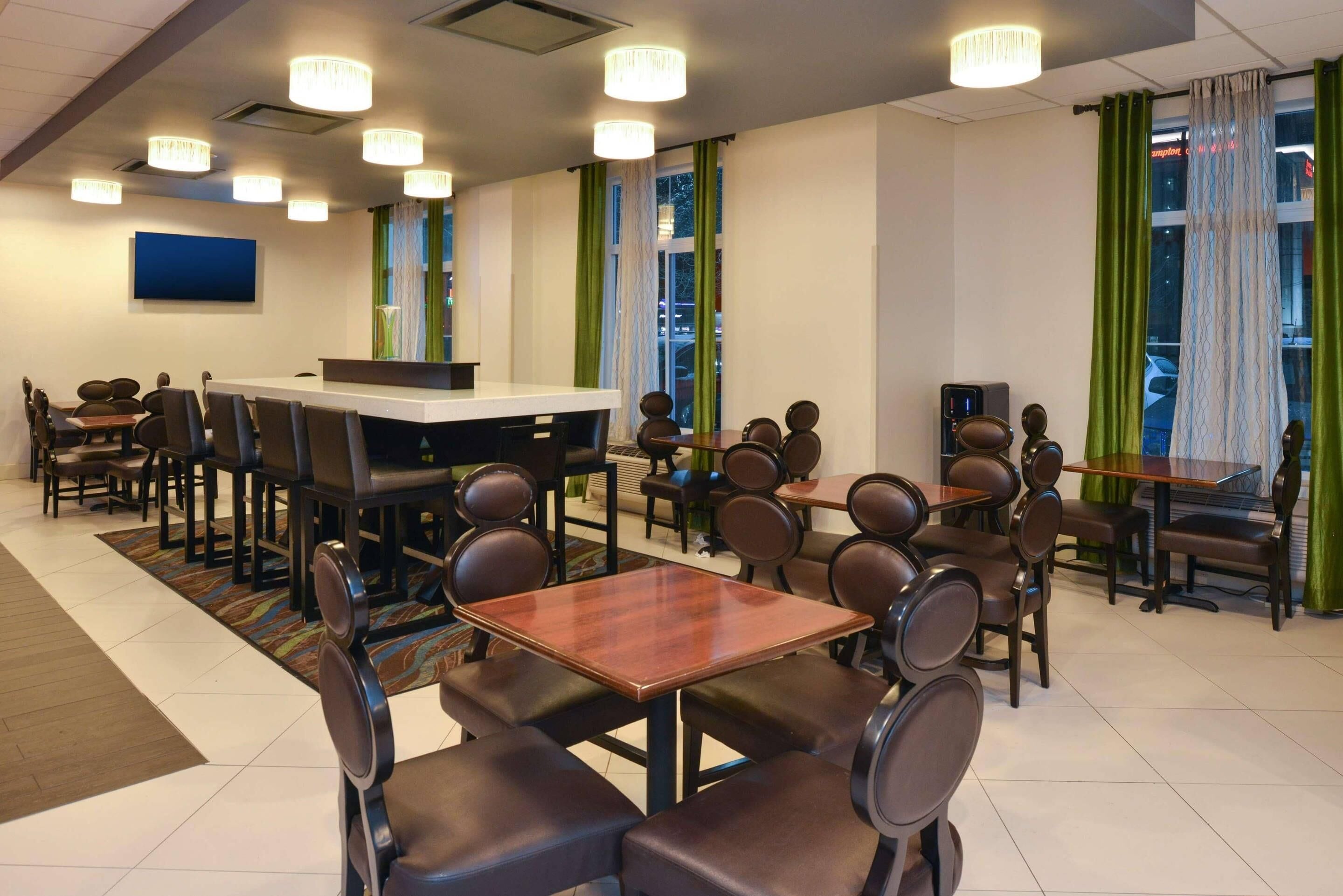 A hotel dining area with multiple tables and chairs, a reception desk, and a large window with curtains.