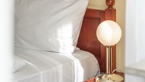 Een netjes opgemaakt bed met witte lakens, een houten hoofdbord en een nachtkastje met een lamp.