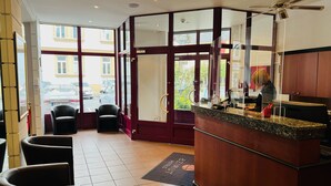 Een hotel lobby met een receptiebalie, een zithoek en grote ramen met uitzicht op de straat.
