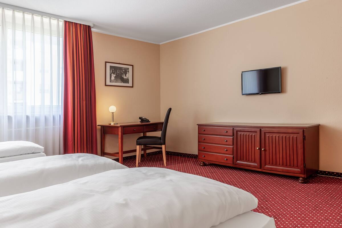 Habitación de hotel con dos camas, un escritorio con silla, cómoda, televisor en la pared y ventana con cortinas.