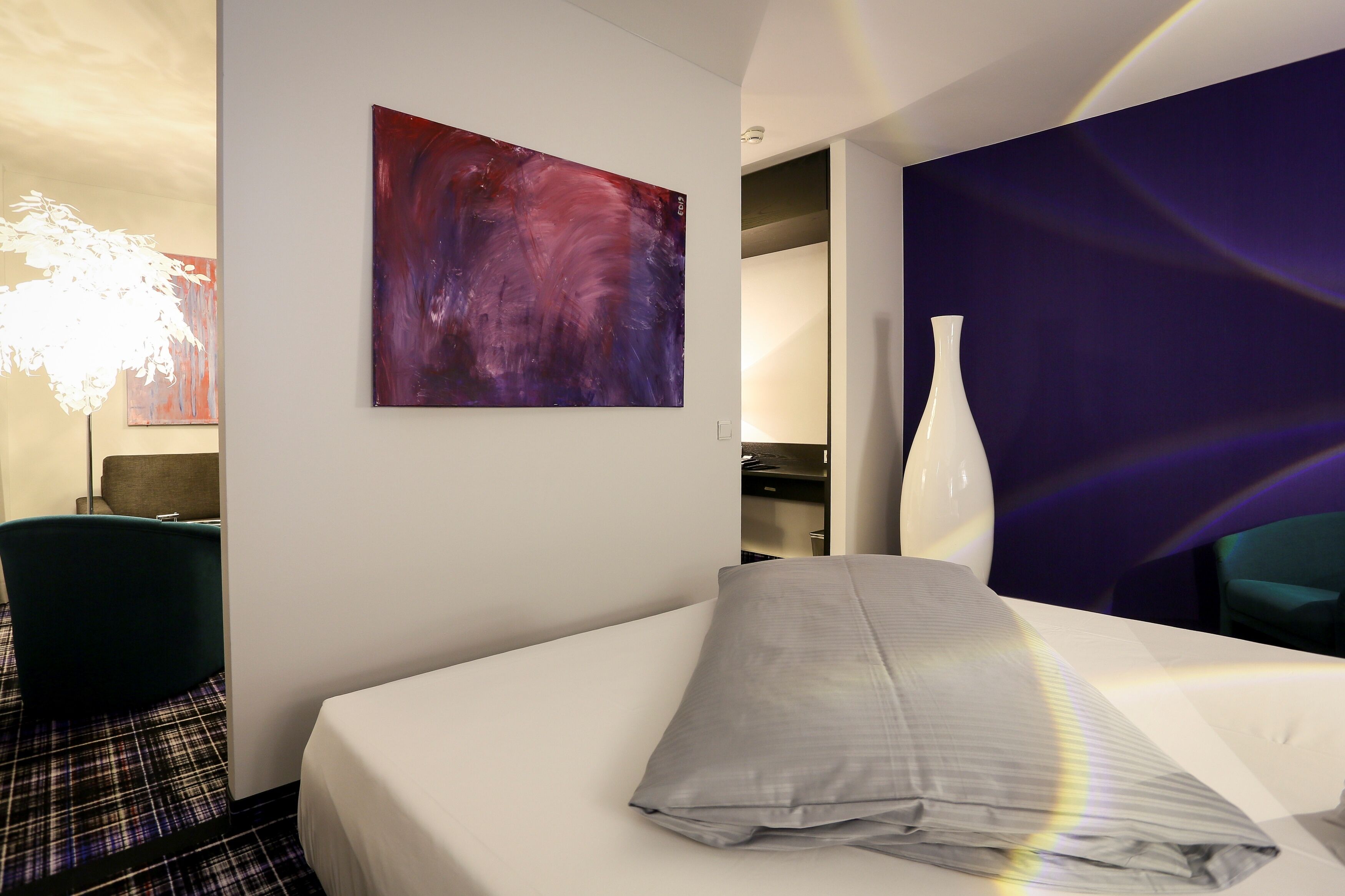 Habitación de hotel con cama, un cuadro abstracto grande, un jarrón blanco y una lámpara moderna.