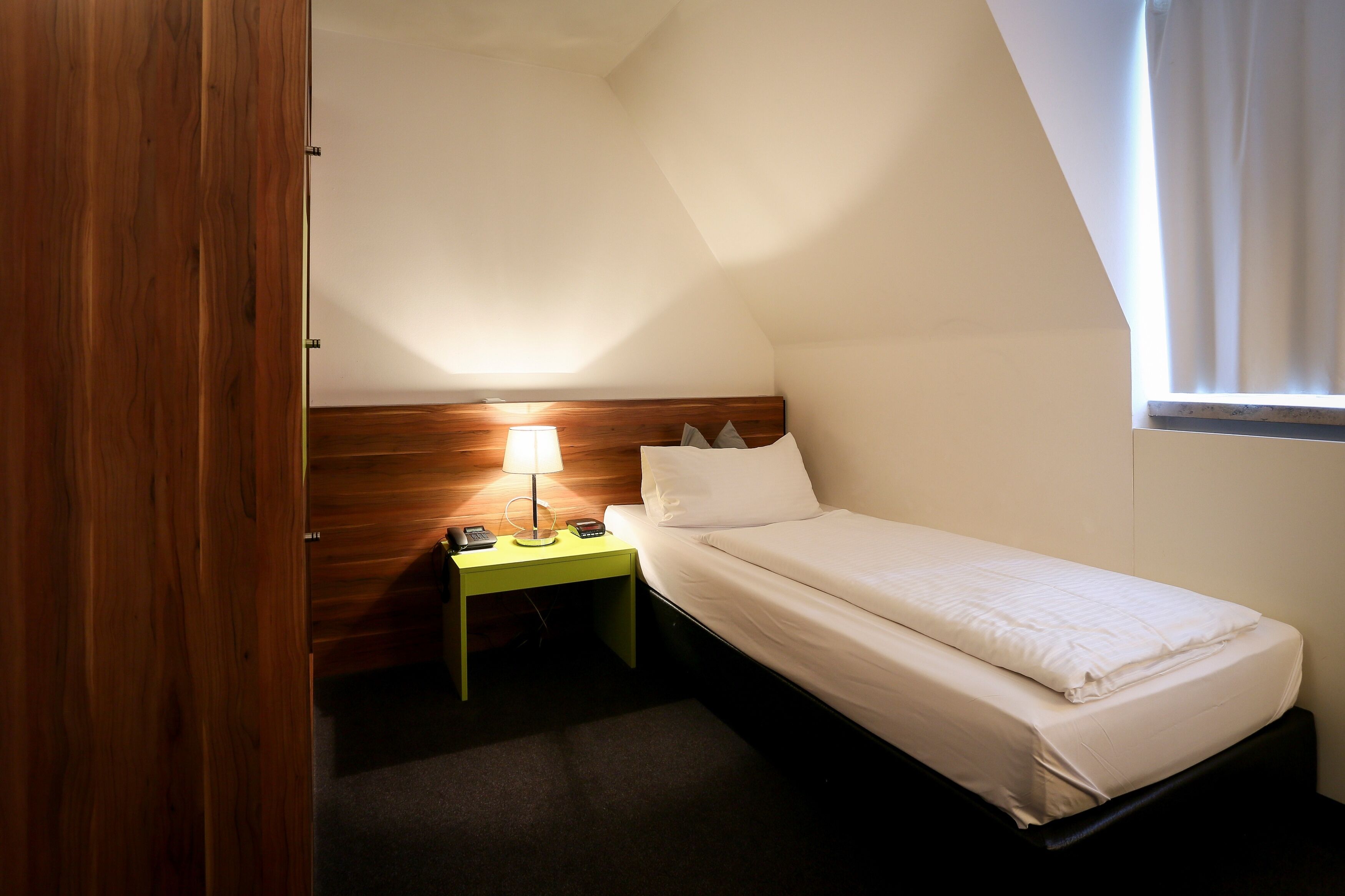 A small, single-bed room with a wooden headboard, a bedside table with a lamp, and a window with curtains.