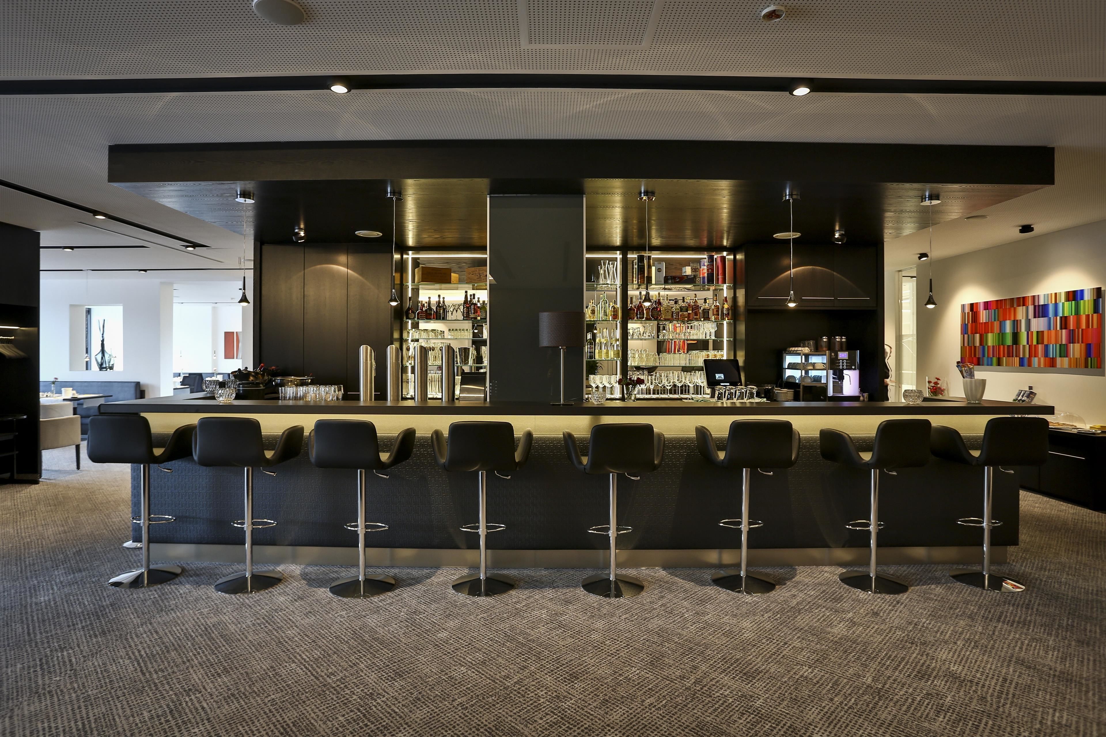 A modern bar area with a long counter, multiple stools, and a variety of bottles and glasses.