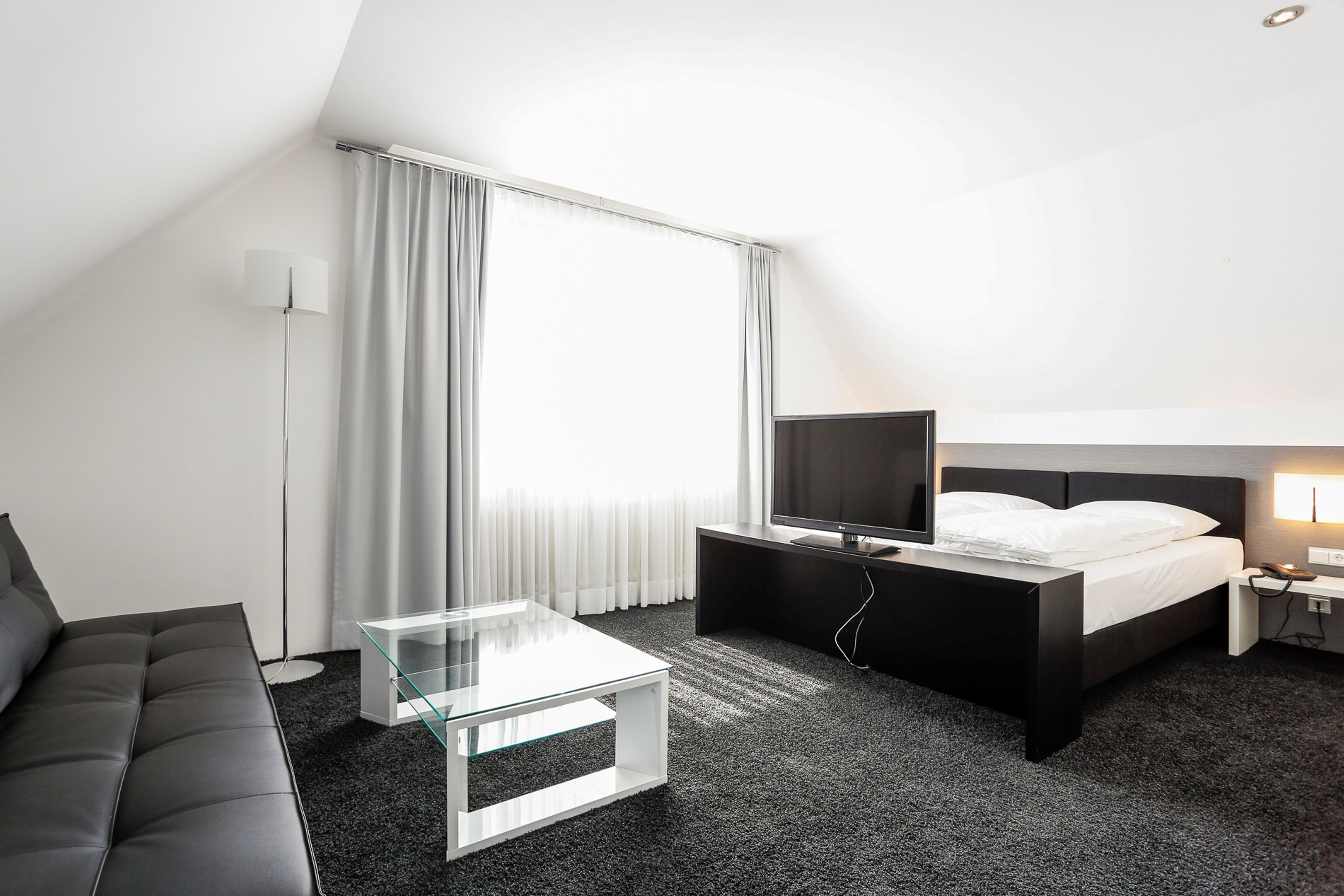 Ein modernes Hotelzimmer mit einer schwarzen Couch, einem Glastisch, einem Flachbildfernseher, einem Bett mit weißer Bettwäsche und einem großen Fenster mit Vorhängen.