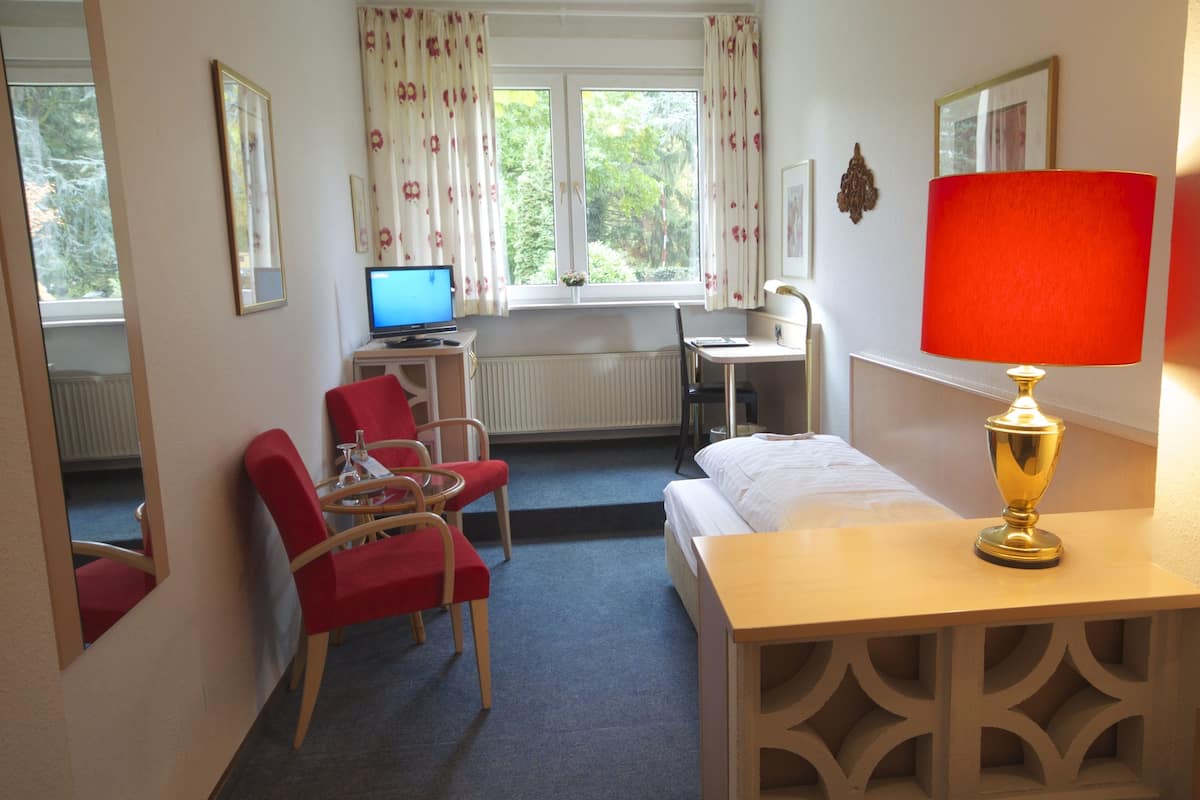 Ein Hotelzimmer mit einem Bett, einem Schreibtisch mit Computer, einem Stuhl, einer Lampe und einem Fenster mit Vorhängen.