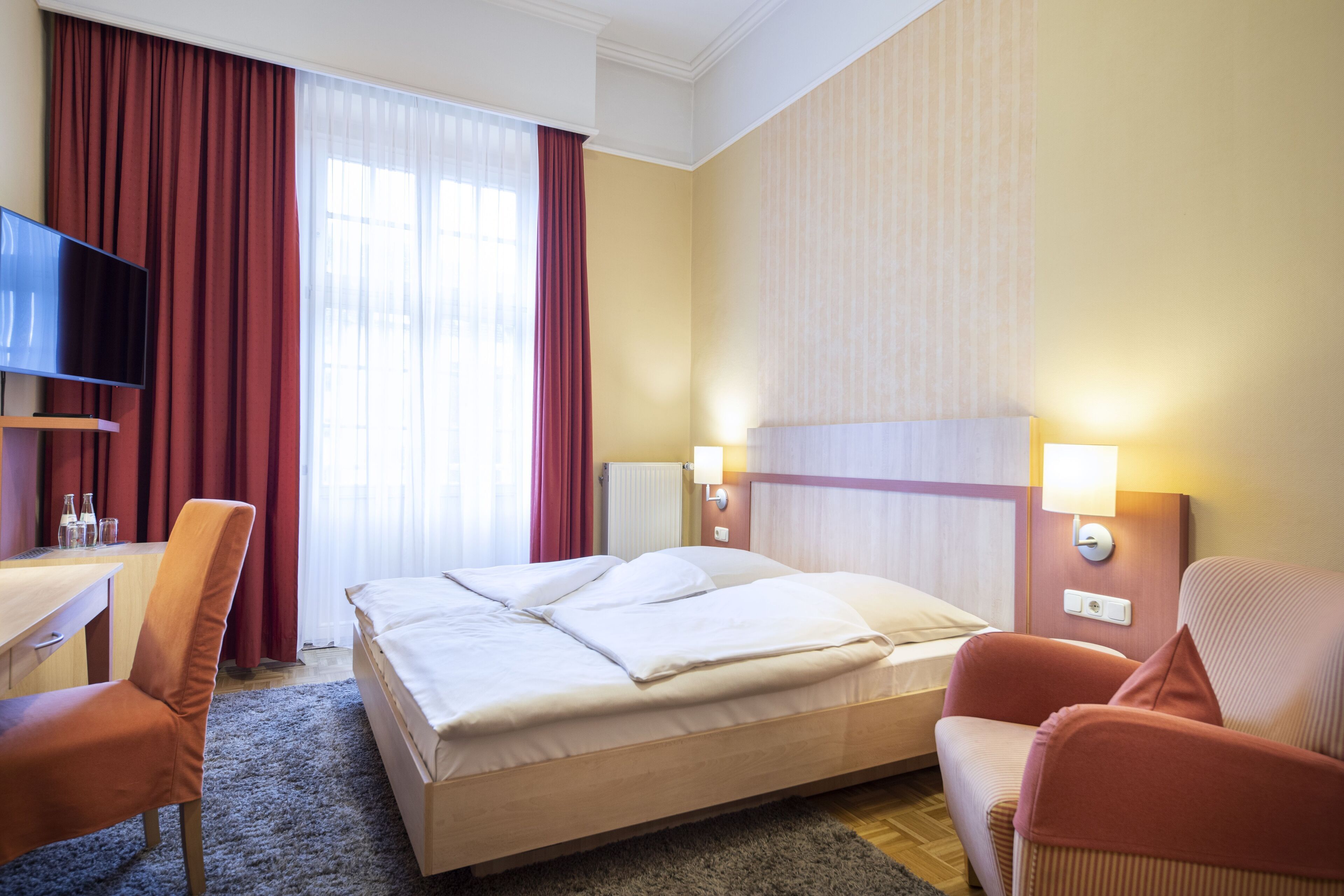 Habitación de hotel con cama, dos sillas, escritorio, televisor y ventana con cortinas.
