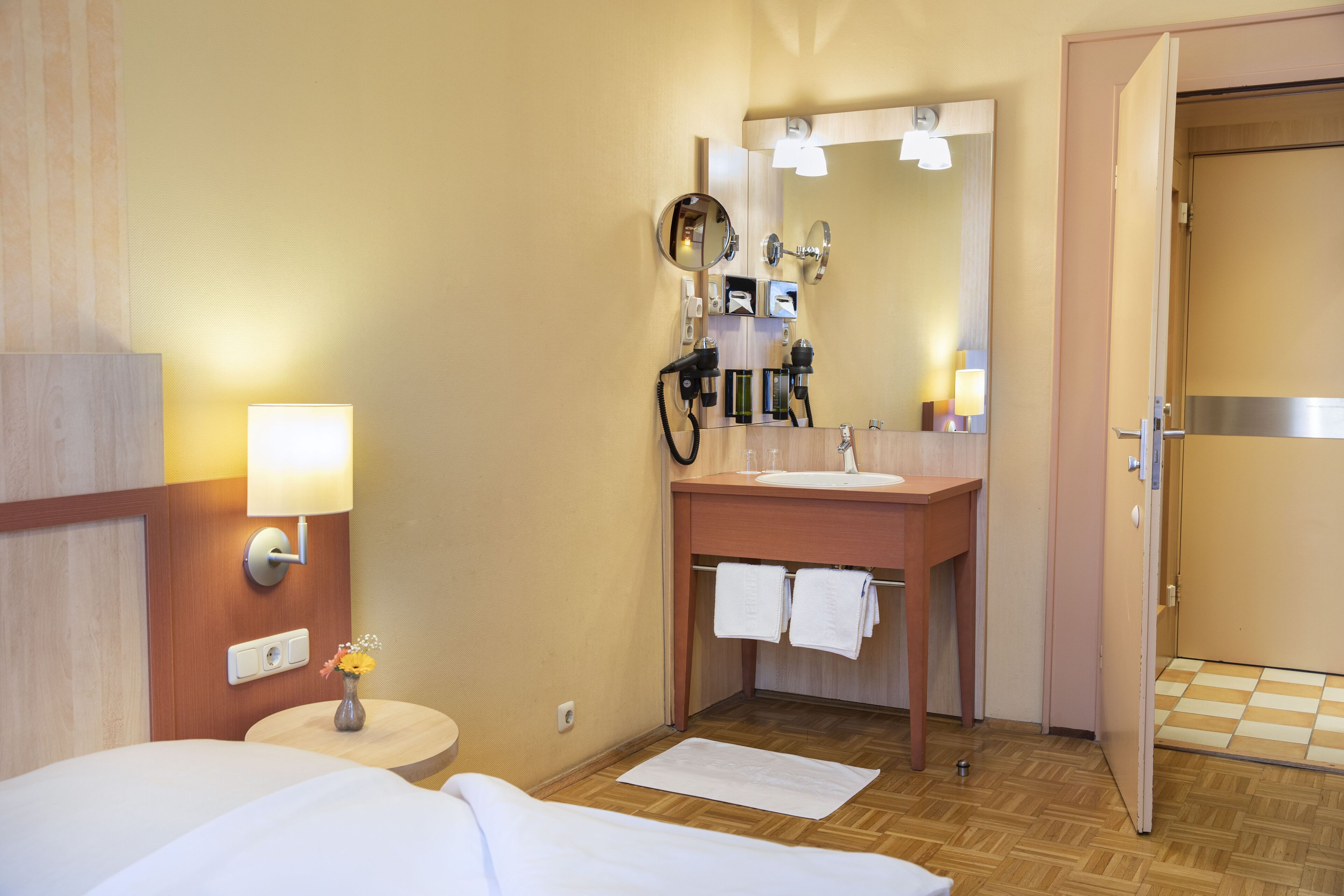 A hotel room with a bed, bedside table, lamp, mirror, and a bathroom with a sink, towel rack, and shower.