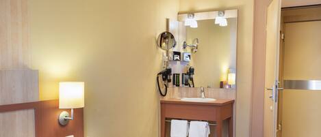 A hotel room with a bed, bedside table, lamp, mirror, and a bathroom with a sink, towel rack, and shower.