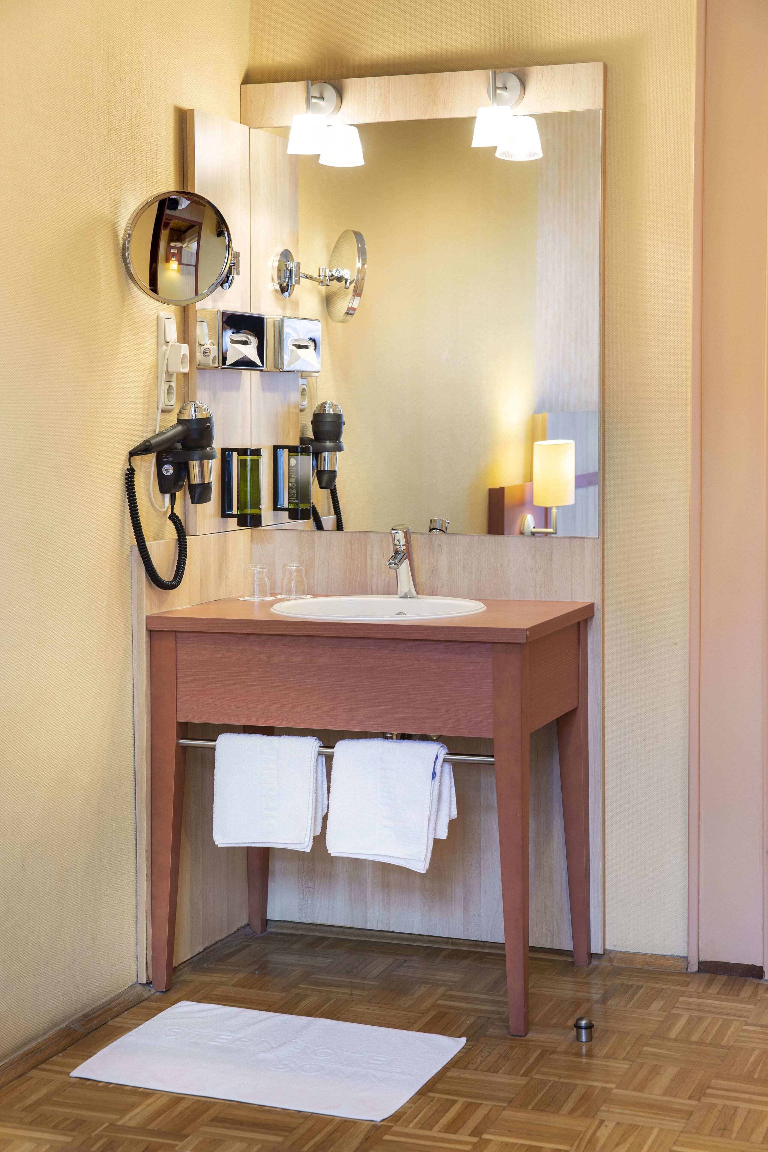 Un baño con tocador de madera, espejo redondo, lavamanos y perchero para toallas.