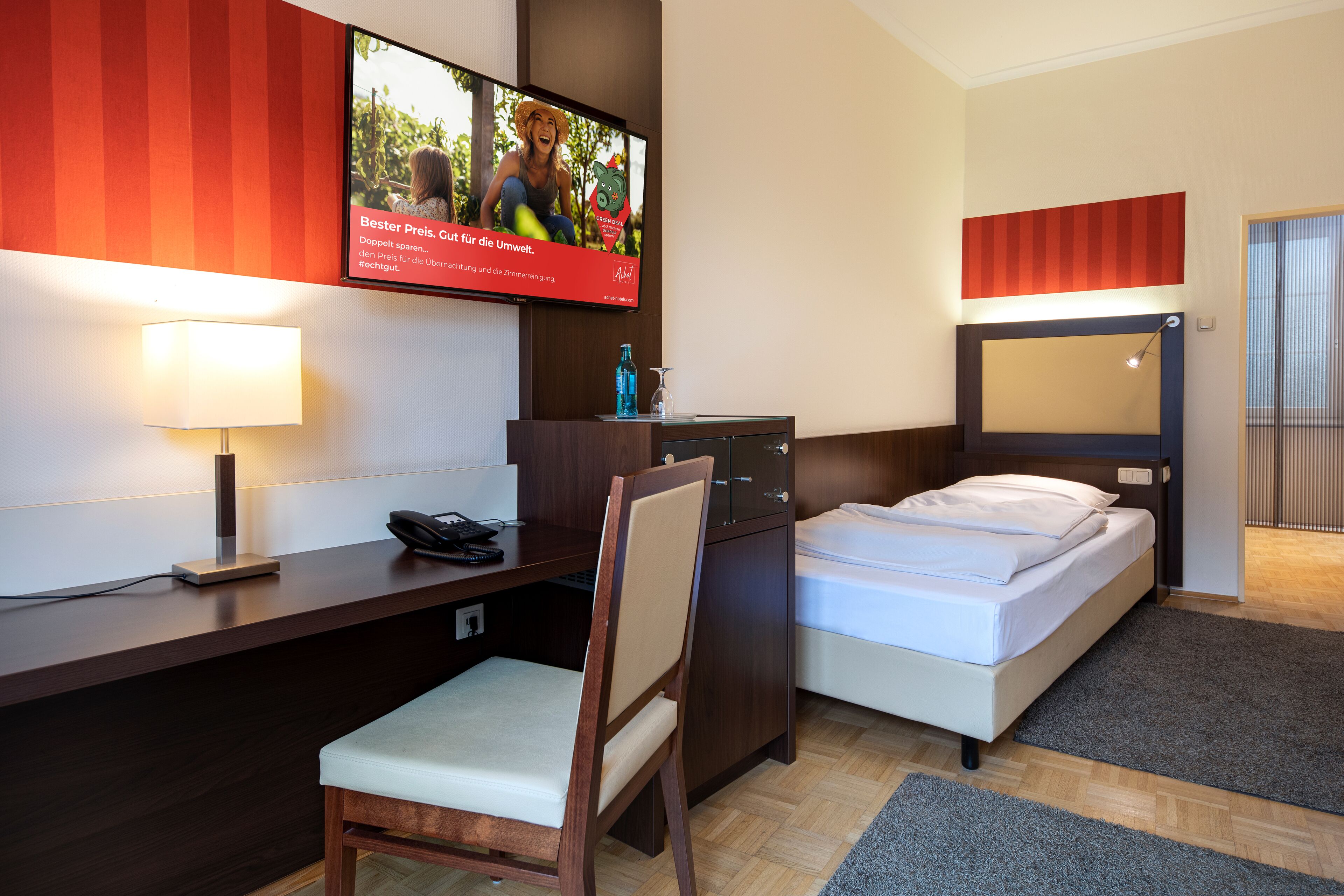 A hotel room with a bed, a desk with a lamp, a chair, a TV mounted on the wall, and a minibar.