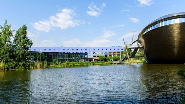 Un hotel moderno con fachada de vidrio, un extenso cuerpo de agua en primer plano y una estructura arquitectónica curva distintiva a la derecha.