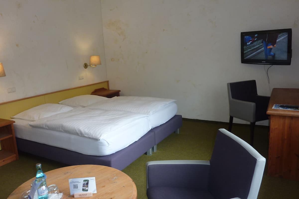 Une chambre d’hôtel équipée d’un lit, d’un bureau, d’une chaise, d’une télévision et d’une petite table sur laquelle se trouvent une bouteille et une carte.