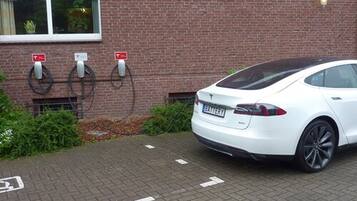 A white Tesla Model S parked at a public charging station with a brick building in the background.