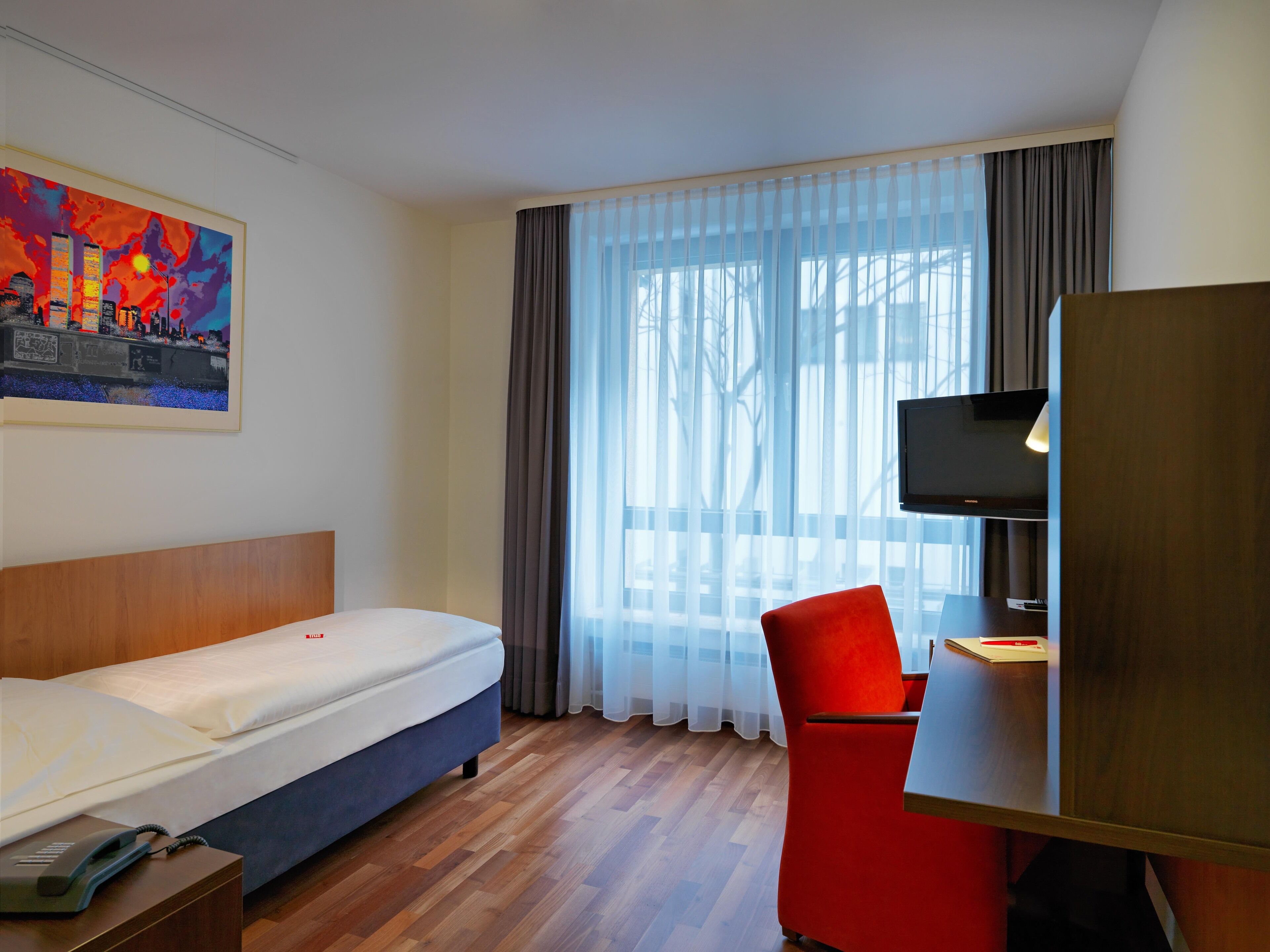 A hotel room with a bed, a desk with a chair, a television, and a painting on the wall.