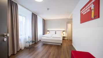 A modern hotel room with a large bed, a small side table, a red bench, and a wall-mounted poster.