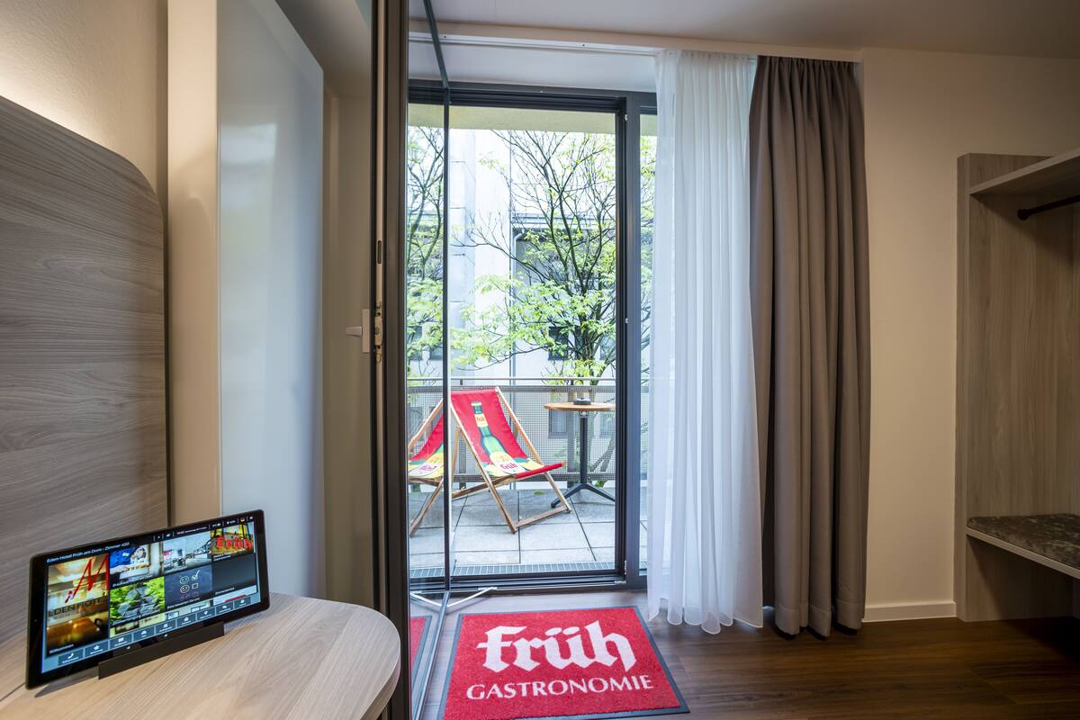 A modern hotel room with a large window, a flat-screen TV, and a red doormat with "Früh Gastronomie" written on it.