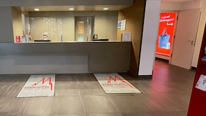 Un espace d’accueil d’hôtel au design moderne, comprenant un comptoir d’accueil, un écran numérique et un panneau de signalisation rouge.