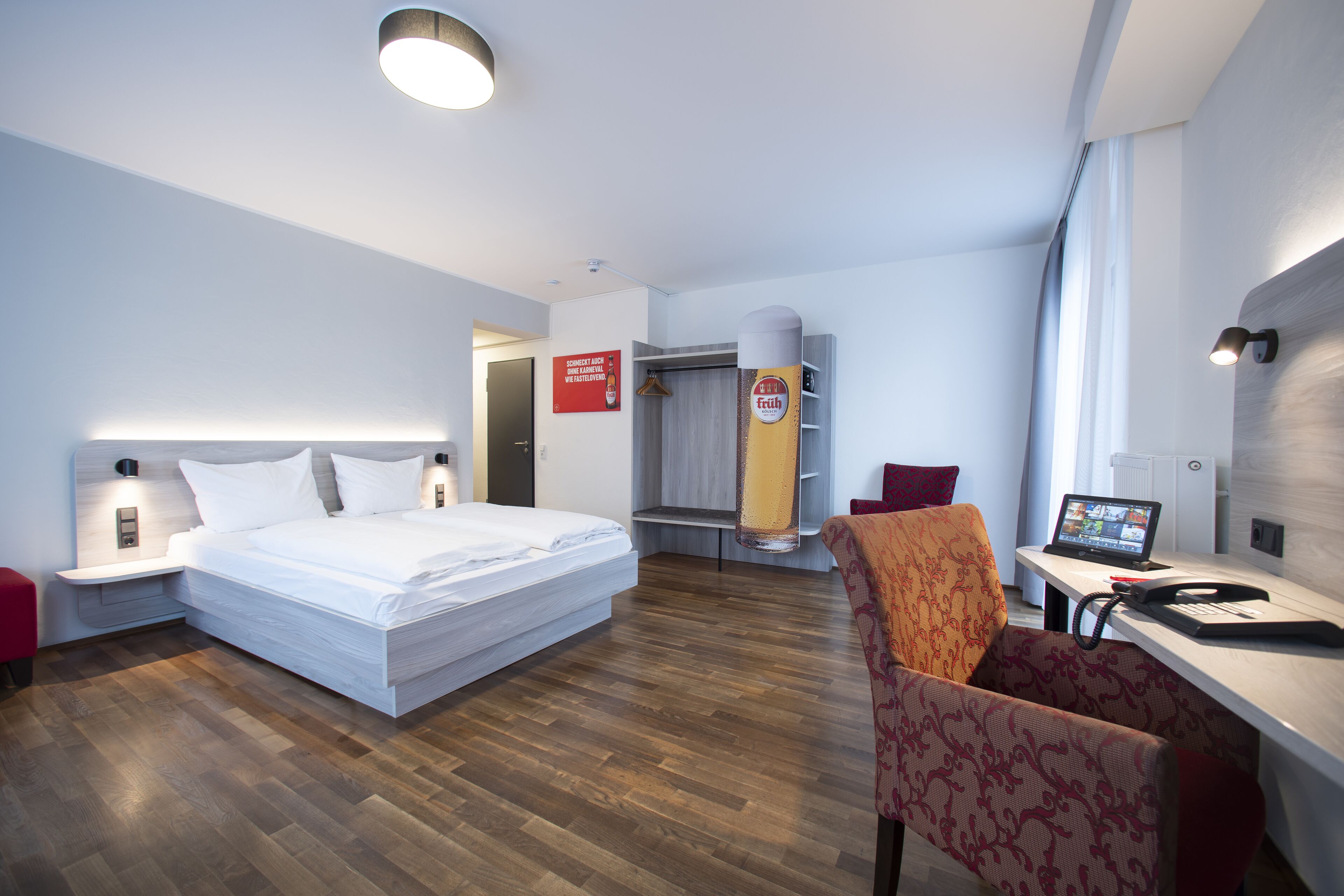 Ein Hotelzimmer mit einem Bett, einem Schreibtisch mit Computer, einem Sessel und einem Duschbereich sowie einem Kleiderschrank.