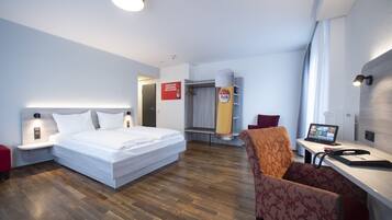 Ein Hotelzimmer mit einem Bett, einem Schreibtisch mit Computer, einem Sessel und einem Duschbereich sowie einem Kleiderschrank.