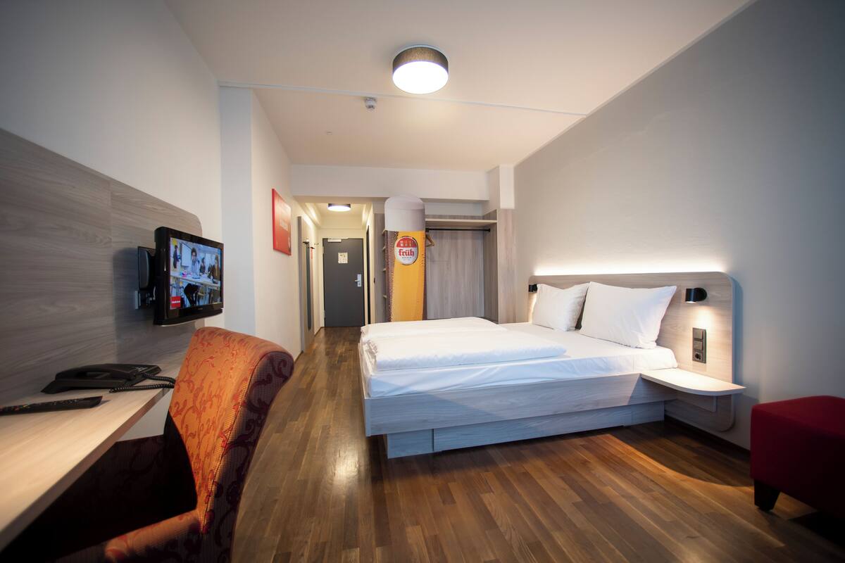 A modern hotel room with a wooden floor, a bed with white linens, a desk with a telephone, and a TV mounted on the wall.