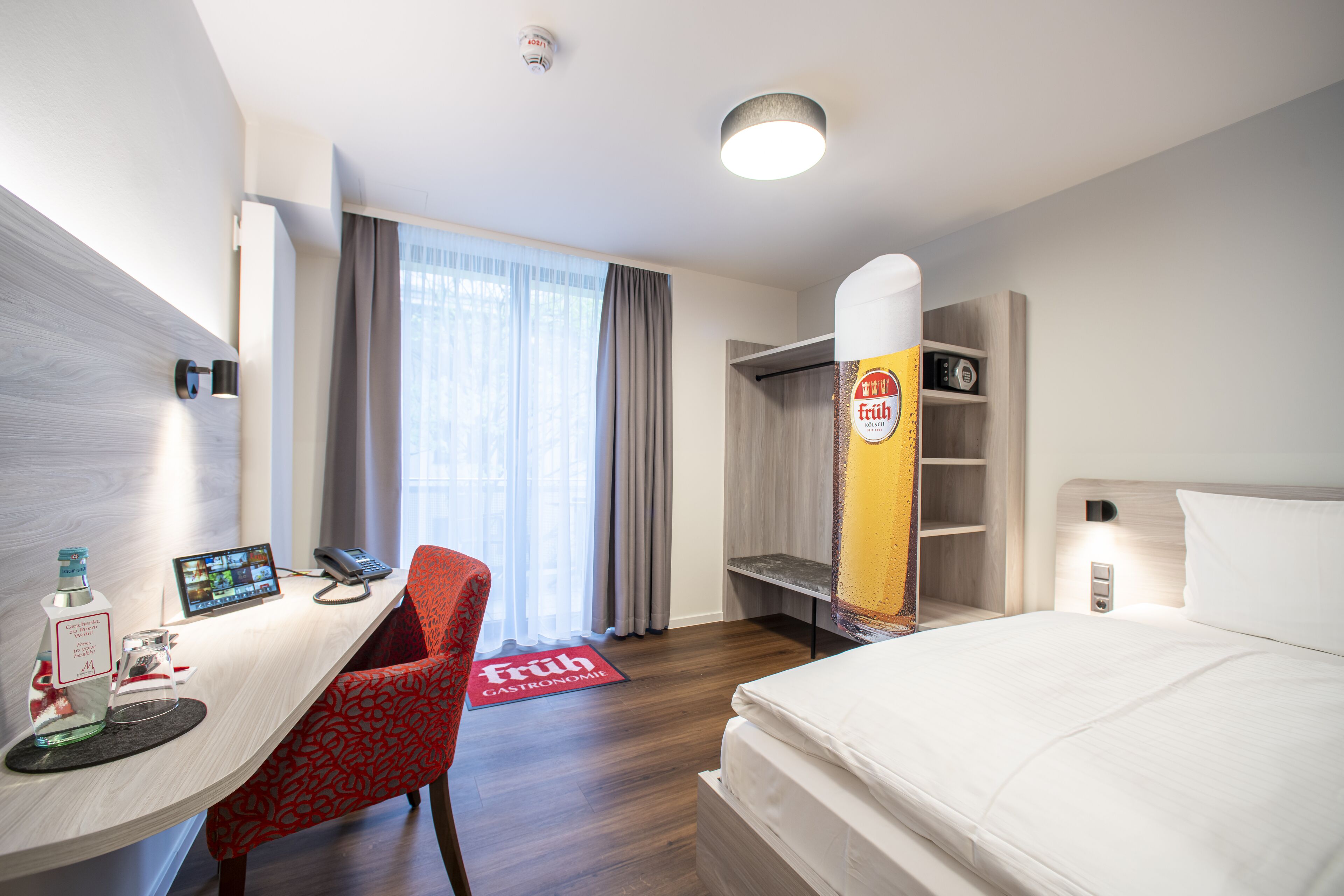 Una habitación de hotel con una cama, un escritorio con teléfono y computadora, una silla roja, un minibar y una repisa para el minibar.